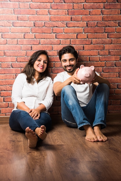 Indian couple with piggy bank sitting in front of red brick background