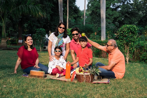 Indian family enjoying picnic - multi generation of asian
family sitting over lawn or green grass in park with fruit basket,
mat and drinks. selective focus