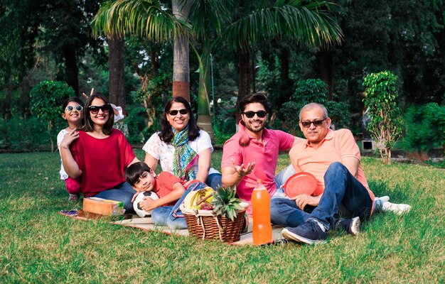 Indian Family enjoying Picnic - Multi generation of asian family sitting over lawn or green grass in park with fruit basket, mat and drinks. selective focus