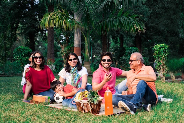 ピクニックを楽しんでいるインドの家族-フルーツバスケット、マット、飲み物を持って公園の芝生や緑の芝生の上に座っているアジアの家族の多世代。セレクティブフォーカス