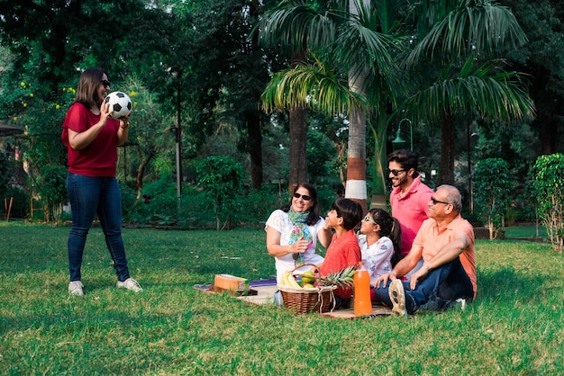 ピクニックを楽しんでいるインドの家族-フルーツバスケット、マット、飲み物を持って公園の芝生や緑の芝生の上に座っているアジアの家族の多世代。セレクティブフォーカス