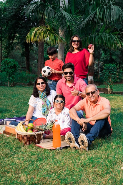 Indian family enjoying picnic - multi generation of asian\
family sitting over lawn or green grass in park with fruit basket,\
mat and drinks. selective focus