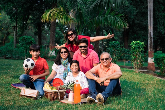 Indian family enjoying picnic - multi generation of asian
family sitting over lawn or green grass in park with fruit basket,
mat and drinks. selective focus