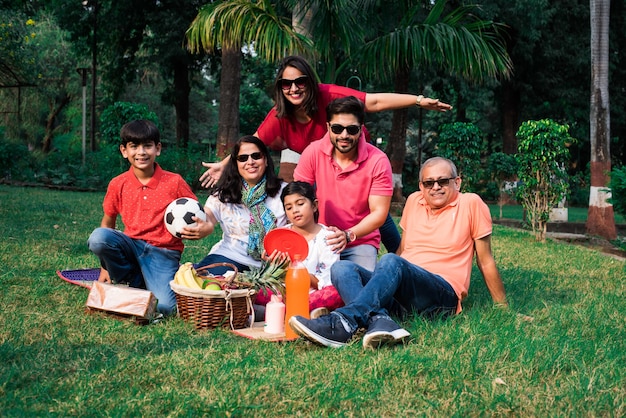 Indian Family enjoying Picnic - Multi generation of asian family sitting over lawn or green grass in park with fruit basket, mat and drinks. selective focus