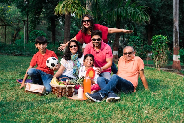 Indian family enjoying picnic - multi generation of asian\
family sitting over lawn or green grass in park with fruit basket,\
mat and drinks. selective focus