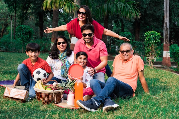 ピクニックを楽しんでいるインドの家族-フルーツバスケット、マット、飲み物を持って公園の芝生や緑の芝生の上に座っているアジアの家族の多世代。セレクティブフォーカス