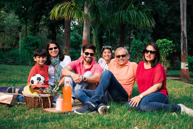 Indian Family enjoying Picnic - Multi generation of asian family sitting over lawn or green grass in park with fruit basket, mat and drinks. selective focus