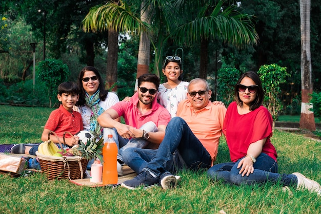 Indian family enjoying picnic - multi generation of asian\
family sitting over lawn or green grass in park with fruit basket,\
mat and drinks. selective focus