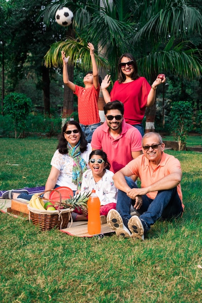 写真 ピクニックを楽しんでいるインドの家族-フルーツバスケット、マット、飲み物を持って公園の芝生や緑の芝生の上に座っているアジアの家族の多世代。セレクティブフォーカス