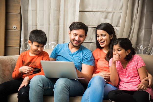 Indian family sitting on sofa and using smartphone laptop or tablet watching movie or surfing internet