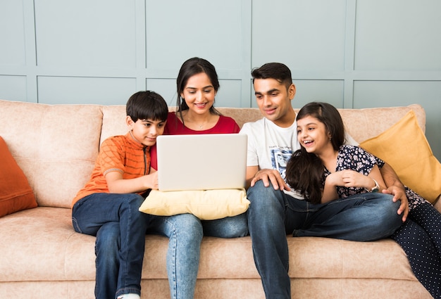 Indian family sitting on sofa and using smartphone, laptop or tablet, watching movie or surfing internet