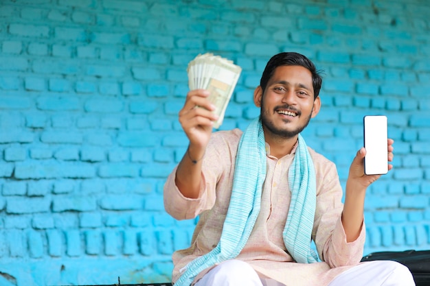 自宅でスマートフォンとお金を見せているインドの農家。
