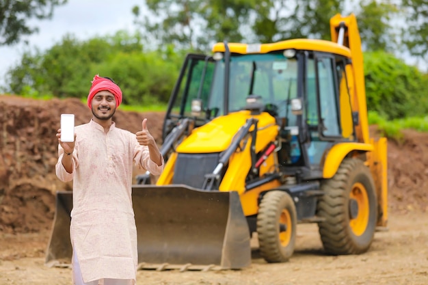 Indian farmer showing smartphone with his new earth mover machine equipment