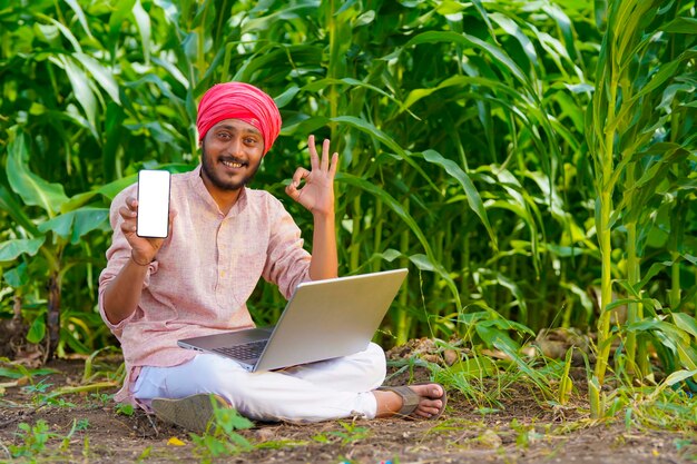 Indian farmer using laptop and showing smartphone at agriculture field.