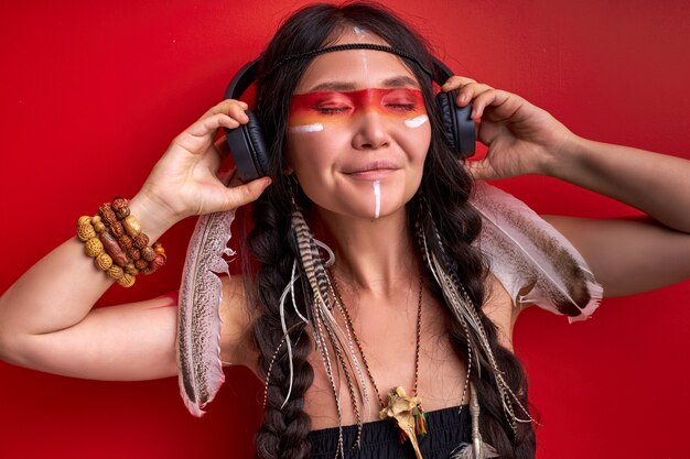 Indian female using headphones, shamanic woman likes music in\
it, listen to music with closed eyes isolated on red wall