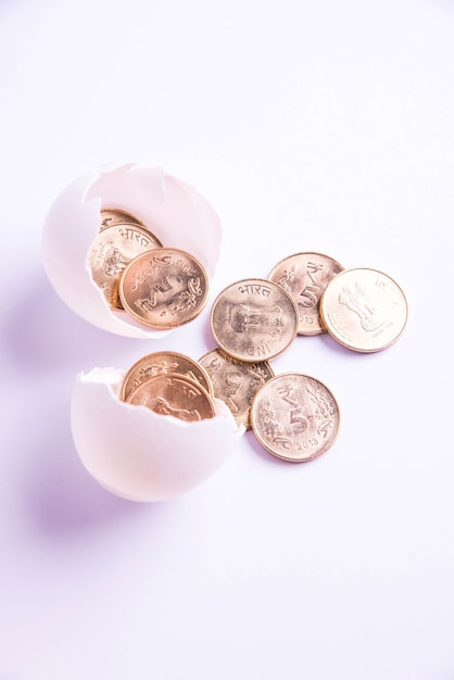 Monete dorate di cinque rupie indiane che emergono dall'uovo incrinato, isolate su sfondo bianco