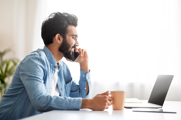 Indian freelancer guy talking on cellphone and drinking coffee at home office