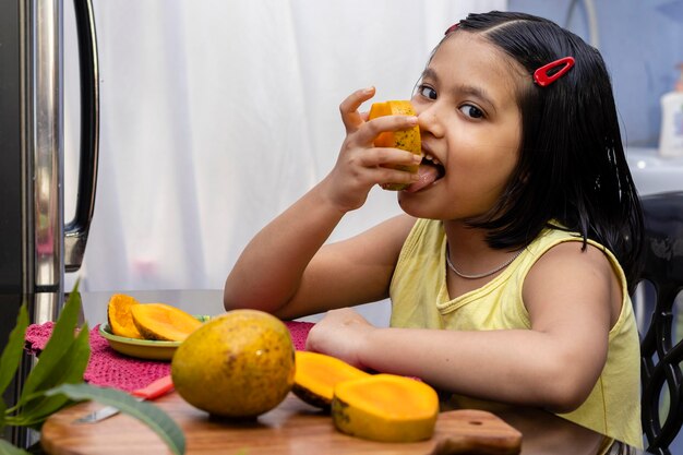 An indian girl child eating mango and smiling at the camera sitting beside a table healthy eating