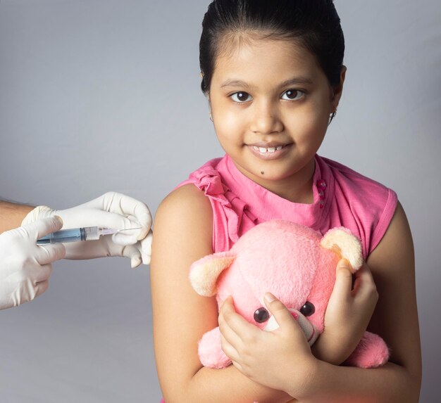 An indian girl child with teddy bear in lap receiving vaccine dose on white background