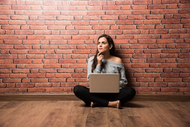 Indian girl or woman sitting with laptop or using laptop over wooden floor against red brick wall