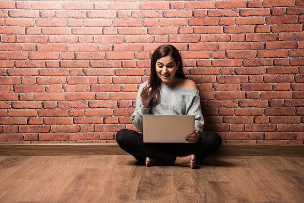 Indian girl or woman sitting with laptop or using laptop over wooden floor against red brick wall