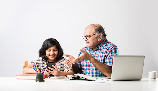 Indian grandfather teaching granddaughter with books, pencil and laptop, home schooling or tuition