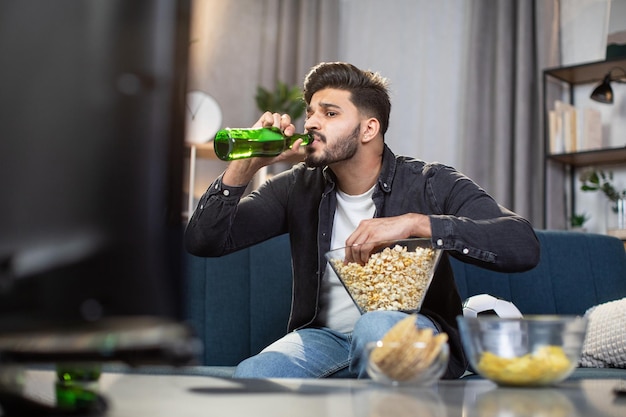 Indian guy drinking beer while watching soccer match at home