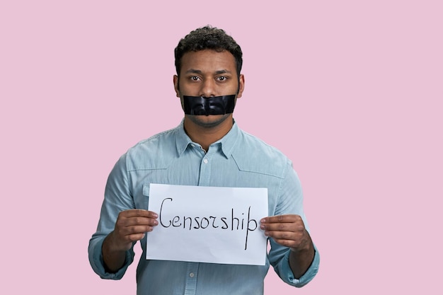 Indian guy silenced with mouth taped holding paper sheet with censorship word isolated on pink backg