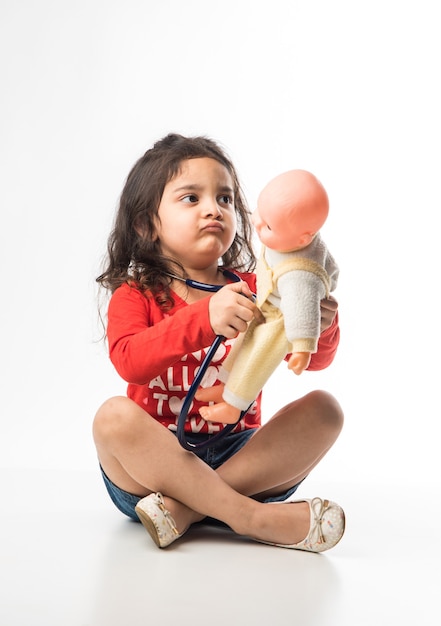 흰색 배경에 앉아 청진기와 박제 아기 또는 강아지 장난감을 가진 인도 어린 소녀