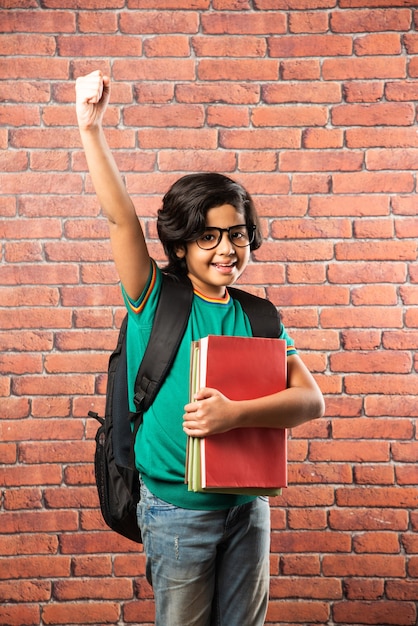 Indian male kid student with spects holding notebooks and school bag against red brick wall