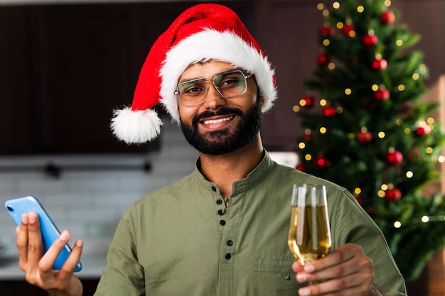 Indian man drinking alcohol at home Christmas tree lights garland background and holding smartphone