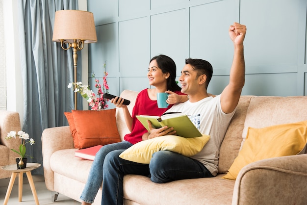 Indian man reading book and wife watching tv while sitting on sofa or couch