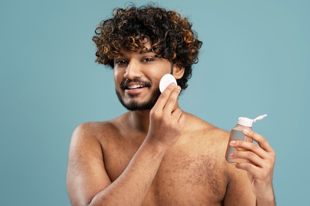 Foto uomo indiano che rimuove il trucco usando un disco di cotone e un tonico per la pelle, pulendo il viso, guardando allo specchio