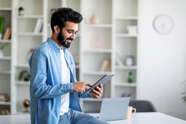 ホームオフィスの机の近くに立ちながらデジタルタブレットを使用するインド人男性