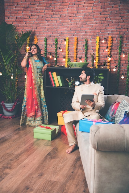 Indian man using tab or tablet touchscreen computer and wife lighting diyas in the background on Diwali festival day