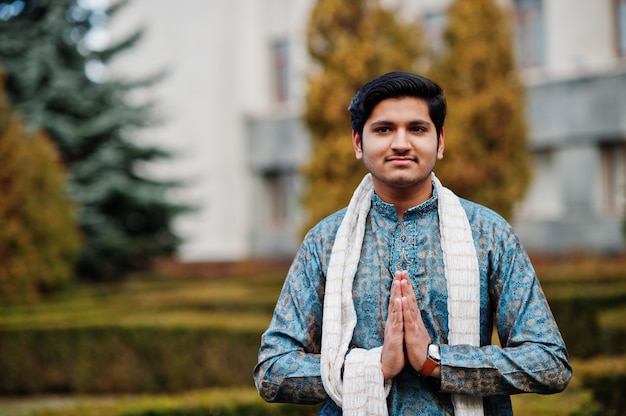 Indian man wear on traditional clothes with white scarf posed outdoor against green bushes at park, show namaste hands sign.