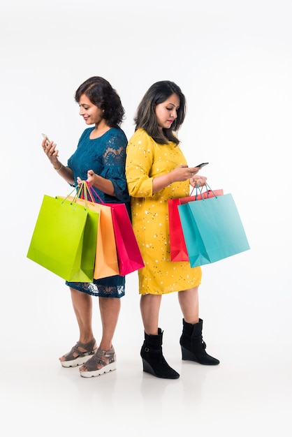 Indian Mother daughter sisters shopping with colourful bags, checking smartphone or taking selfie while standing isolated over white background