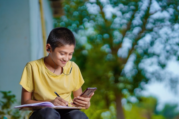 Indian school boy holding phone distance learning class using mobile application, watching online lesson, video calling in app making notes studying at home