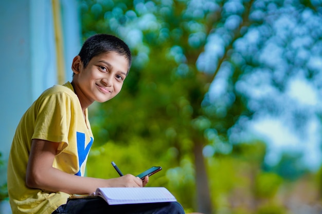 Indian school boy holding phone distance learning class using mobile application, watching online lesson, video calling in app making notes studying at home