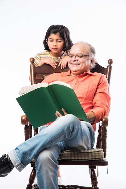 Indian Senior man or grandpa reading book with granddaughter while sitting on rocking chair against white wall