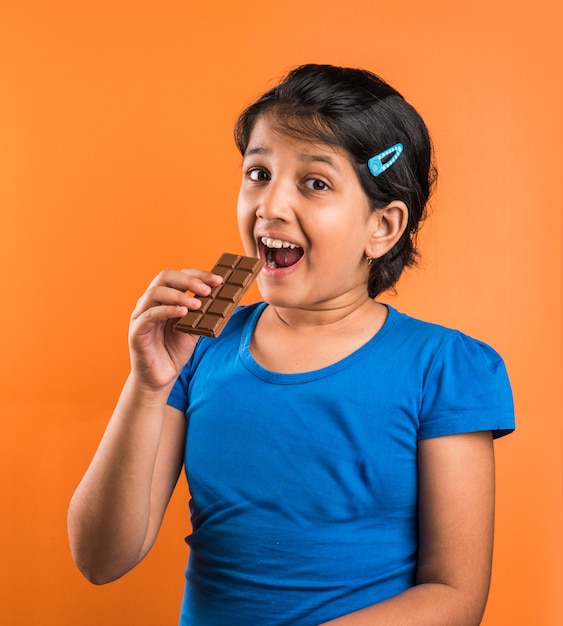 オレンジ色の背景で隔離のチョコレートスラブを食べるインドの小さな女の子