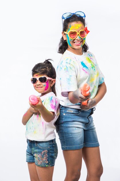 Indian small kids or friends or siblings celebrating Holi festival with gulal or powder colour, sweets, pichkari or spray, isolated over white background