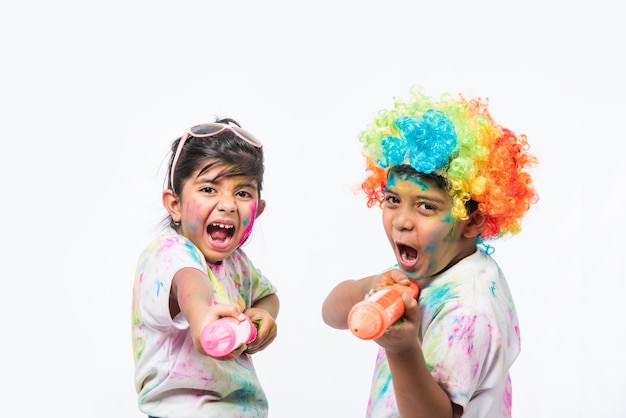 Indian small kids or friends or siblings celebrating Holi festival with gulal or powder colour, sweets, pichkari or spray, isolated over white background