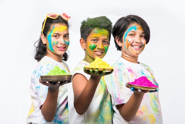 Indian small kids or friends or siblings celebrating Holi festival with gulal or powder colour, sweets, pichkari or spray, isolated over white background