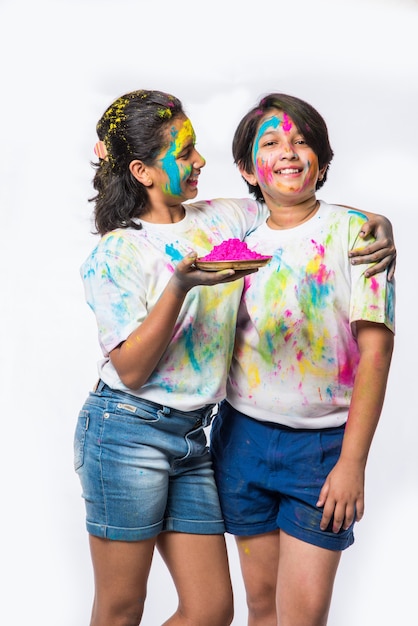 Indian small kids or friends or siblings celebrating Holi festival with gulal or powder colour, sweets, pichkari or spray, isolated over white background