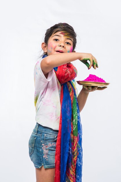 Indian small kids or friends or siblings celebrating Holi festival with gulal or powder colour, sweets, pichkari or spray, isolated over white background