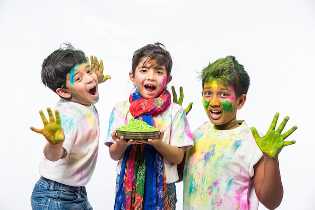 Indian small kids or friends or siblings celebrating Holi festival with gulal or powder colour, sweets, pichkari or spray, isolated over white background