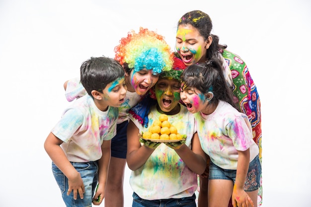 Indian small kids or friends or siblings celebrating Holi festival with gulal or powder colour, sweets, pichkari or spray, isolated over white background