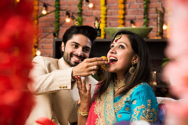 Indian smart couple eating sweet laddu on Diwali or anniversary, selective focus