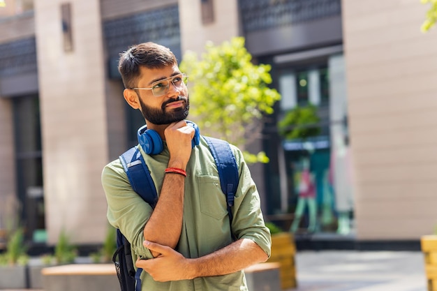 Studente indiano con auricolare blu e zaino che sembra pensieroso in una giornata di sole